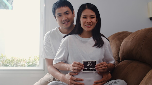 젊은 아시아 임신 몇 표시 및 뱃속에서 초음파 사진 아기를 찾고. 엄마와 아빠가 집에서 거실에서 소파에 누워있는 아이를 돌보는 동안 평화로운 미소가 행복하다고 느낍니다.