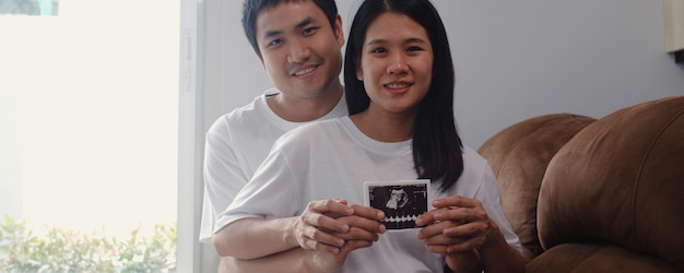 若いアジアの妊娠カップルを表示し、お腹の中で超音波写真の赤ちゃんを探しています。ママとパパは、自宅のリビングルームのソファに横たわっている子供の世話をしながら平和な幸せな笑顔を感じています。