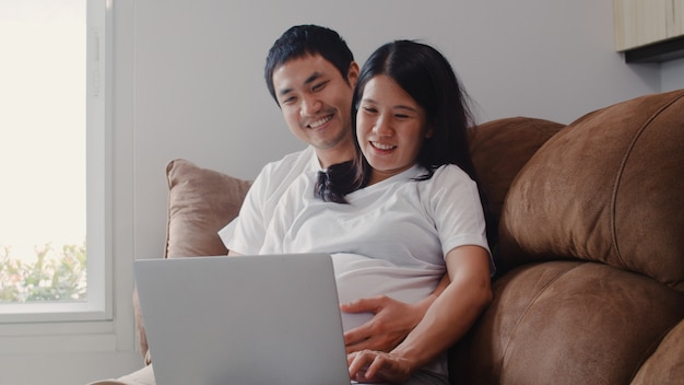 노트북 검색 임신 정보를 사용하여 젊은 아시아 임신 부부. 엄마와 아빠가 집에서 거실에서 소파에 누워있는 그들의 아이를 돌보는 동안 긍정적이고 평화로운 미소 행복을 느끼고 있습니다.