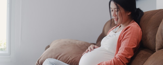Молодая азиатская беременная женщина держа ее живот разговаривая с ее ребенком. Мама чувствует себя счастливым, улыбаясь позитивным и мирным, пока заботиться о ребенке, беременность лежа на диване в гостиной дома