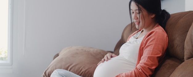 Young Asian Pregnant woman holding her belly talking with her child. Mom feeling happy smiling positive and peaceful while take care baby, pregnancy lying on sofa in living room at home .