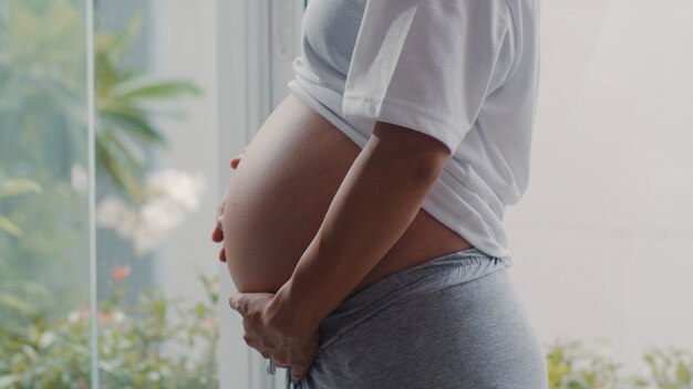 젊은 아시아 임신 한 여자는 그녀의 아이와 이야기하는 그녀의 배꼽을 잡고. 엄마 돌보는 아기, 임신 집에서 창문 가까이 임신 동안 긍정적이 고 평화로운 미소 행복.