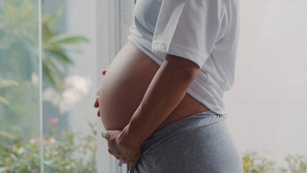 Young Asian Pregnant woman holding her belly talking with her child. Mom feeling happy smiling positive and peaceful while take care baby, pregnancy near window in living room at home .