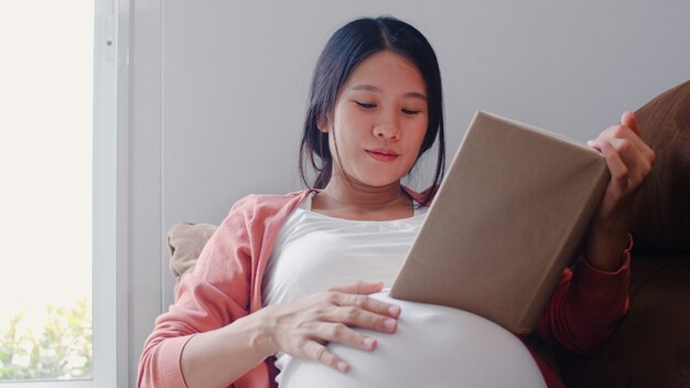 Young Asian Pregnant woman read a book for baby in belly. Mom feeling happy smiling positive and peaceful while take care child lying on sofa in living room at home .