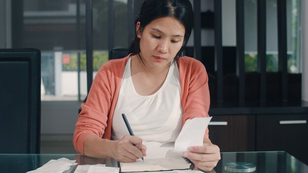 若いアジア妊婦の自宅での収入と支出の記録。ラップトップの記録予算、税、財務書類、自宅のリビングルームで働くeコマースを使用して幸せなママの女の子。