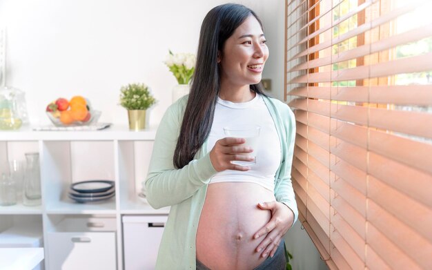 출산 직전까지 임신 중 건강을 유지하기 위해 신선한 우유 한 잔을 들고 부엌 방에 서 있는 젊은 아시아 임산부