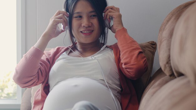 Young Asian Pregnant woman using phone and headphone play music for baby in belly. Mom feeling happy smiling positive and peaceful while take care child lying on sofa in living room at home .