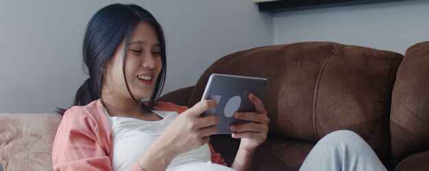 タブレット検索妊娠情報を使用して若いアジアの妊娠中の女性。自宅のリビングルームのソファーに横たわっている彼女の子供の世話をしながらママは前向きで平和な笑顔幸せを感じています。