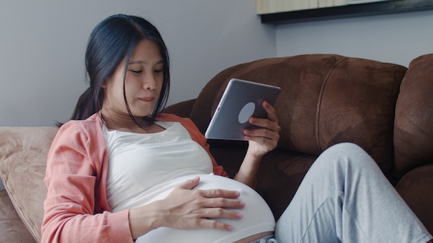 Young Asian Pregnant woman using tablet search pregnancy information. Mom feeling happy smiling positive and peaceful while take care her child lying on sofa in living room at home .