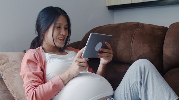 태블릿 검색 임신 정보를 사용하여 젊은 아시아 임신 여자. 엄마 집에서 거실에서 소파에 누워 그녀의 아이를 돌보는 동안 긍정적이고 평화로운 미소 행복 느낌.