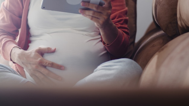 Free photo young asian pregnant woman using tablet search pregnancy information. mom feeling happy smiling positive and peaceful while take care her child lying on sofa in living room at home .