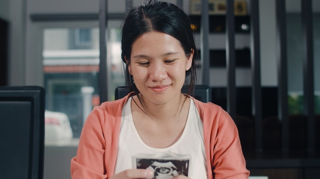 Le giovani donne incinte asiatiche mostrano e guardano il bambino della foto di ultrasuono in pancia. mamma che ritiene felice sorridente pacifico mentre abbi cura del bambino che si siede sulla tavola in salone a casa di mattina.