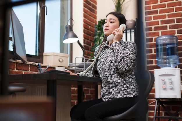 Young asian project manager talking on landline with marketing team leader about uneffective promoting campaign. Advertising company office agent being informed by telephone about work policy.