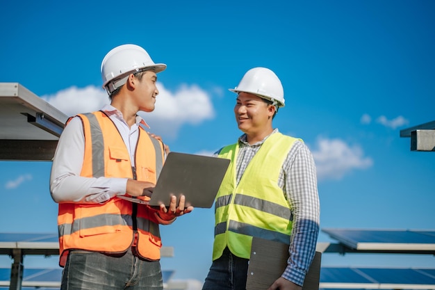 Young Asian technician man and colleague in safety uniform checking operation of sun and photovoltaic solar panel and use laptop computer while working in solar farm