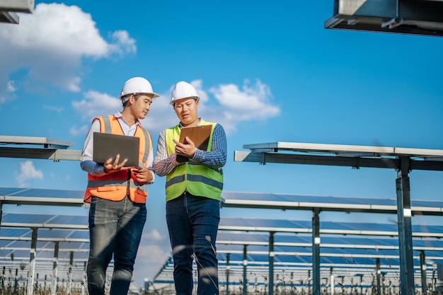 Giovane tecnico asiatico e collega in uniforme di sicurezza che controllano il funzionamento del sole e del pannello solare fotovoltaico e utilizzano il computer portatile mentre si lavora nella fattoria solare