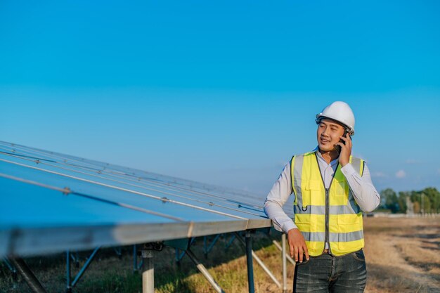 太陽光発電ソーラーパネルのコピースペースの長い列の間に立ってスマートフォンで話している若いアジアの技術者の男