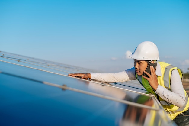 Young Asian technician man standing and talking on smartphone between long rows of photovoltaic solar panels copy space