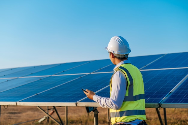 Young Asian technician man standing and talking on smartphone between long rows of photovoltaic solar panels copy space