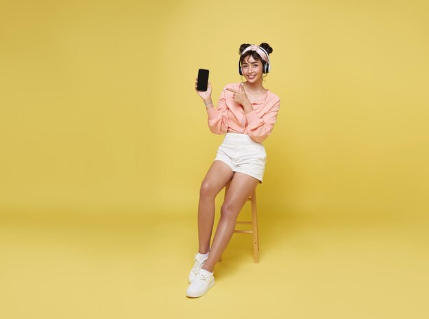 Young asian teenage girl hand pointing smart phone While her listening music in headphones and sitting on chair isolate on yellow background