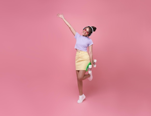 Young asian teenage girl holding skateboard with wearing wireless headphones listening to music