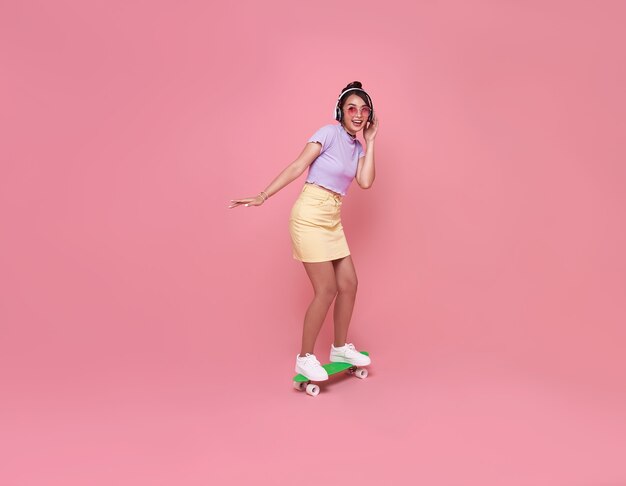 Молодая азиатская девочка-подросток играет на скейтборде с носить беспроводные наушники, слушая музыку на розовой стене.