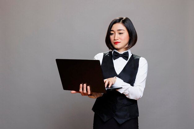 Young asian waitress using laptop, checking cafe online menu and managing catering service. Attractive restaurant receptionist wearing professional uniform and holding portable computer in studio