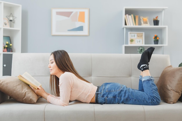 Молодая азиатка в повседневной одежде лежит на диване у себя дома и читает книгу, счастливая и позитивная, расслабляющая, проводящая выходные дома
