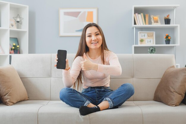 カジュアルな服を着た若いアジア人女性が自宅のソファに座ってスマートフォンを持って親指を立てて笑顔で幸せで前向きな時間を家で過ごす