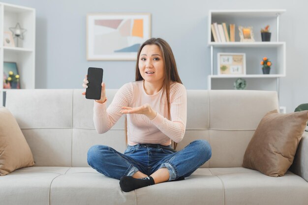 молодая азиатская женщина в повседневной одежде сидит на диване в домашнем интерьере, представляя смартфон, недовольна и сбита с толку, проводя время дома