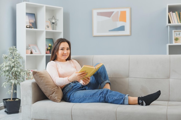Молодая азиатка в повседневной одежде сидит на диване у себя дома и читает книгу, счастливая и позитивная, расслабляющая, проводящая выходные дома