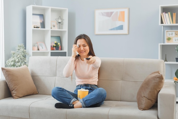 Young asian woman in casual clothes sitting on a couch at home interior with bucket of popcorn holding remote watching television being sad crying rubbing eye with tissue