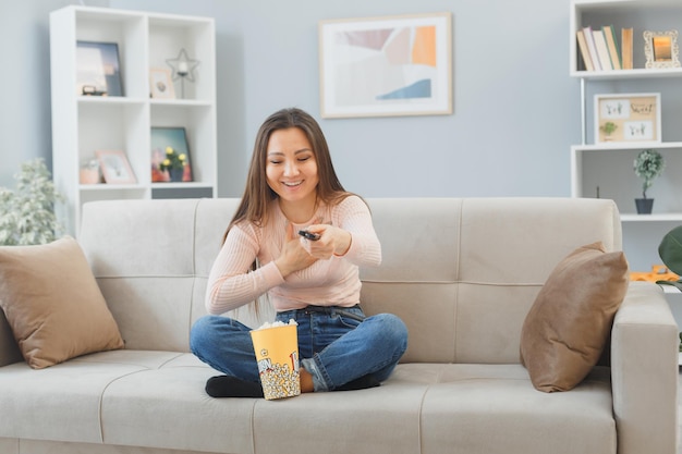 Giovane donna asiatica in abiti casual seduto su un divano a casa interna con secchio di popcorn che tiene la televisione a distanza guardando felice e positivo sorridente allegramente