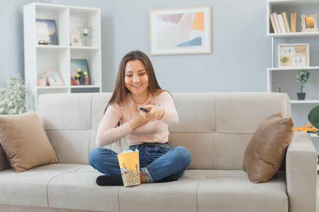Молодая азиатка в повседневной одежде сидит на диване в домашнем интерьере с ведром попкорна, держа в руках пульт дистанционного управления, смотрит телевизор, счастливая и позитивная, весело улыбаясь