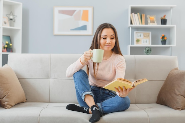 Молодая азиатская женщина в повседневной одежде сидит на диване в домашнем интерьере с кружкой чая и читает книгу, счастливая и позитивная, расслабляющая, проводящая выходные дома