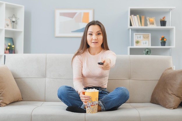 Giovane donna asiatica in abiti casual seduto su un divano a casa con secchio di popcorn che tiene in mano guardare la televisione a distanza felice e positivo sorridente amichevole trascorrere del tempo a casa