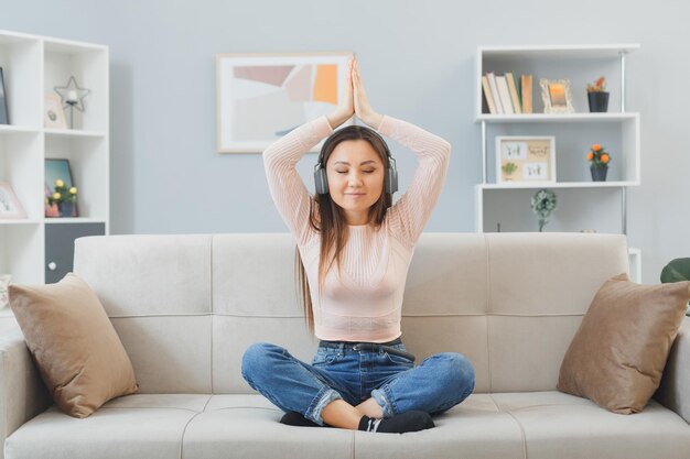カジュアルな服を着た若いアジア人女性が自宅のソファに座ってヘッドフォンを持って幸せで前向きにリラックスして手のひらを頭に抱えて瞑想する