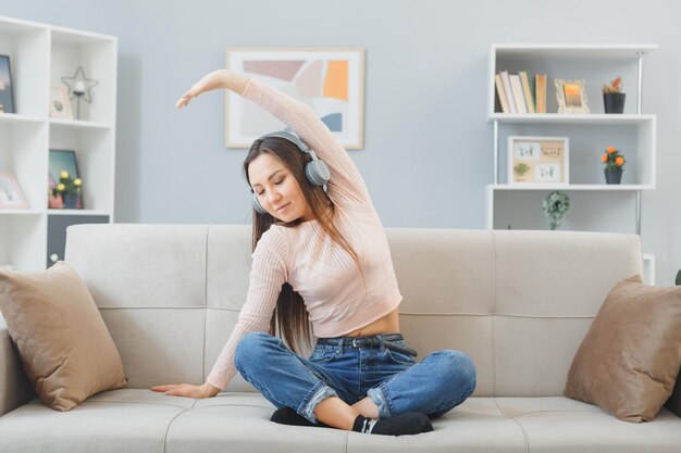 캐주얼 옷을 입은 젊은 아시아 여성, 헤드폰을 끼고 집 내부 소파에 앉아 행복하고 긍정적인 휴식을 취하며 자신을 스트레칭