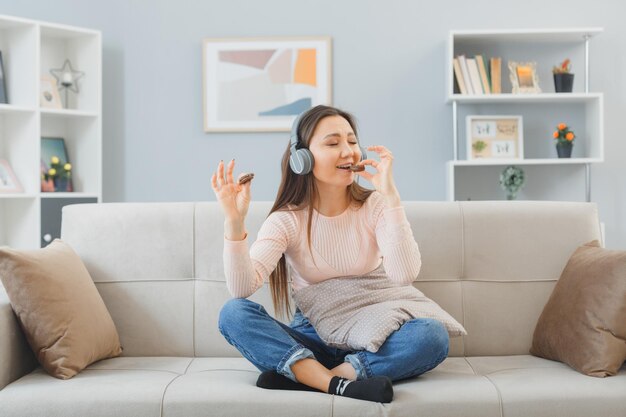 Young asian woman in casual clothes with headphones sitting on a couch at home interior holding remote watching television eating cookies having fun