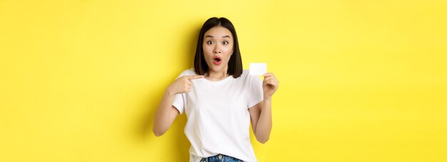 カジュアルな白い t シャツを着た若いアジア女性プラスチック製のクレジット カードを示し、黄色のカメラで笑顔