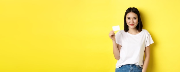 Free photo young asian woman in casual white tshirt showing plastic credit card and smiling at camera yellow ba