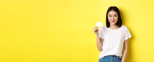 Young asian woman in casual white tshirt showing plastic credit card and smiling at camera yellow ba