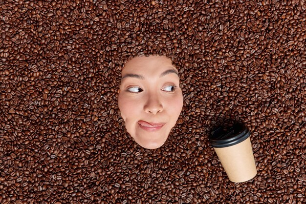 Молодая азиатская любительница кофе смотрит на аппетитную чашку освежающего напитка, облизывая губы языком, окруженным коричневыми жареными семечками, содержащими большое количество антиоксидантов