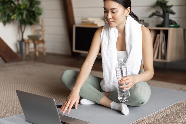 Young asian woman connect to online fitness instructor on laptop workout via video chat at home sitt...