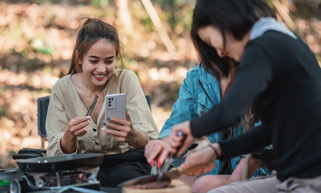 요리를 하는 젊은 아시아 여성과 그녀의 친구는 팬에서 식사를 하는 것을 즐깁니다. 그들은 자연 공원에서 캠핑하는 동안 함께 즐겁게 이야기하고 웃는다