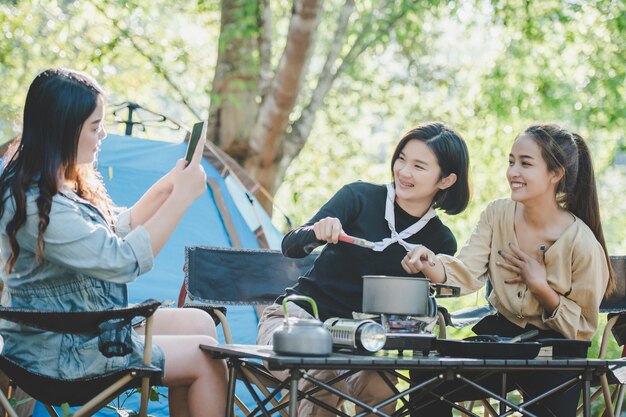 요리를 하는 젊은 아시아 여성과 그녀의 친구는 냄비에 식사를 만드는 것을 즐깁니다. 그들은 자연 공원에서 캠핑하는 동안 함께 즐겁게 이야기하고 웃는다