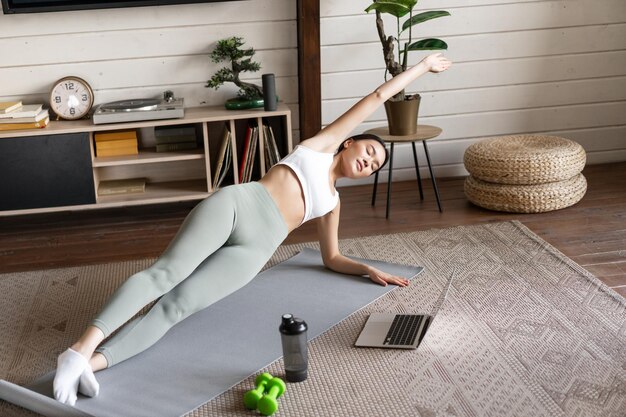 Young asian woman doing workout at home on floor mat following sport instructor on laptop video stre...