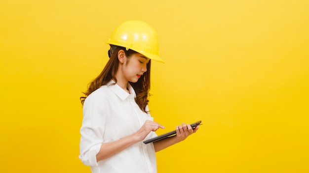 Young Asian woman engineer using digital tablet with positive expression, dressed in casual clothing and looking at camera over yellow wall. Happy adorable glad woman rejoices success.