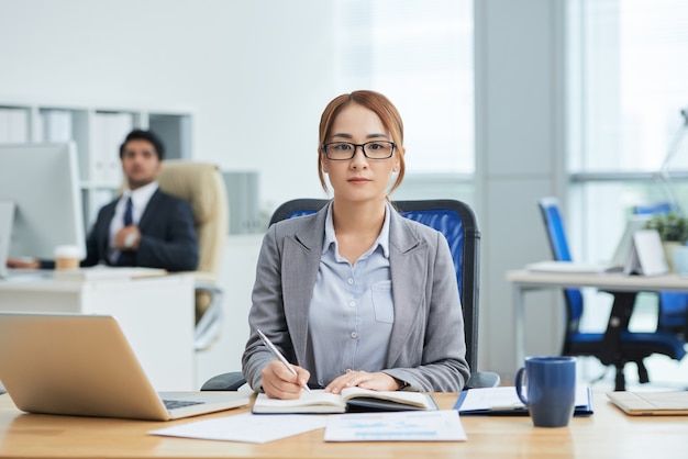 Young asian woman in glasses sitting at desk in office, writing in planner and looking at camera