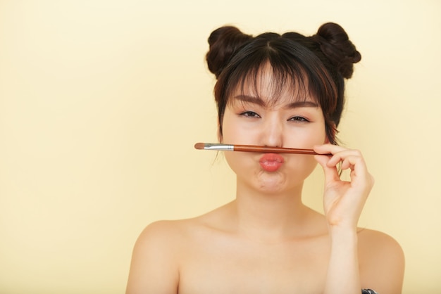 Free photo young asian woman grimacing with eyeshadow brush between her lips and nose