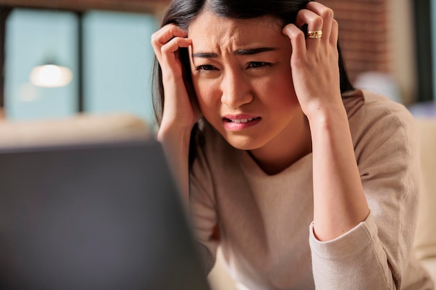 Young asian woman having migraine and headache while trying to work from home. Tired female on couch covering forehead and exhausted red eyes unhappy sick upset ill fever depresion